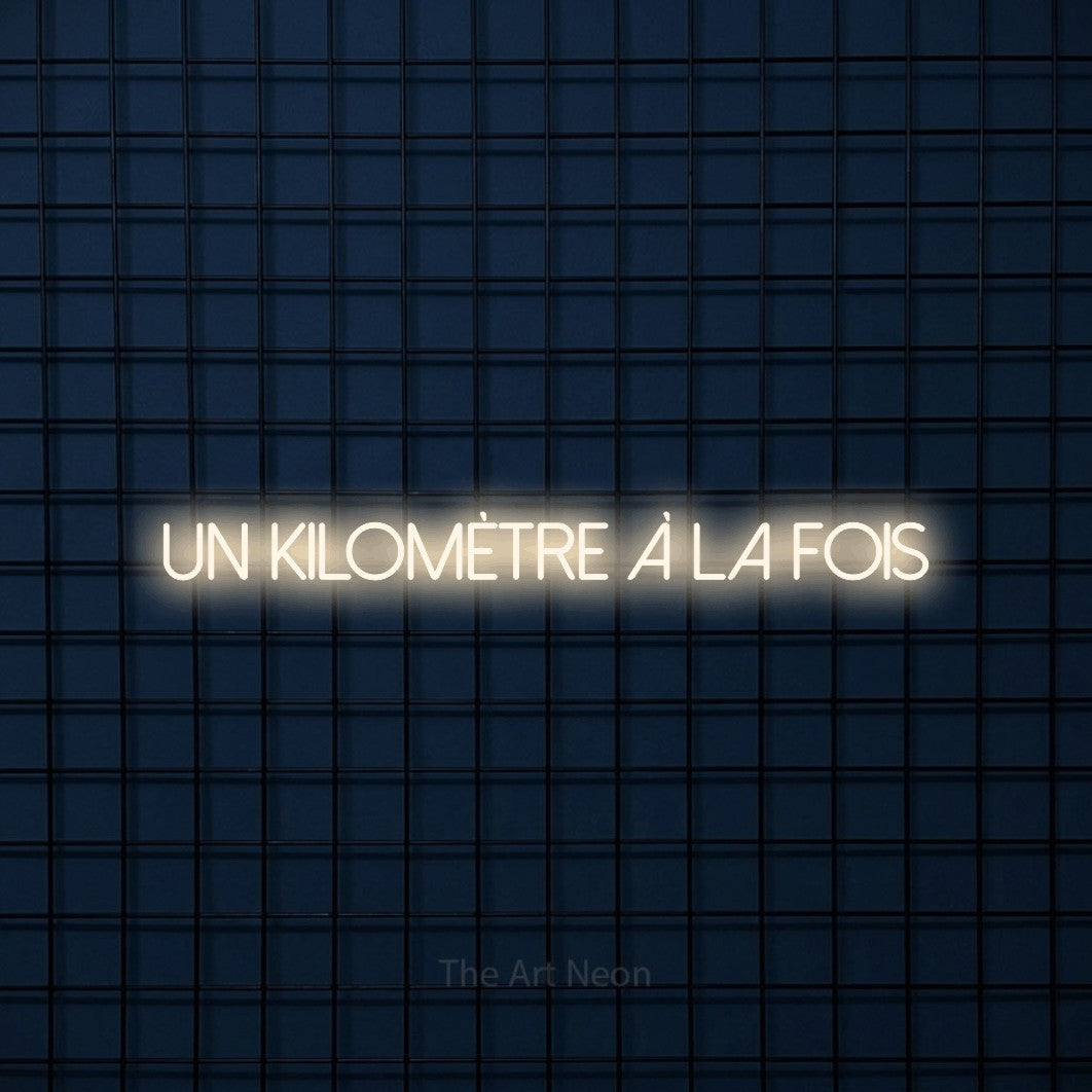 Néon LED « Un kilomètre à la fois » en blanc chaud, en lettres fines sans empattement, mis en scène sur une grille bleu foncé, pour un espace d’entraînement.