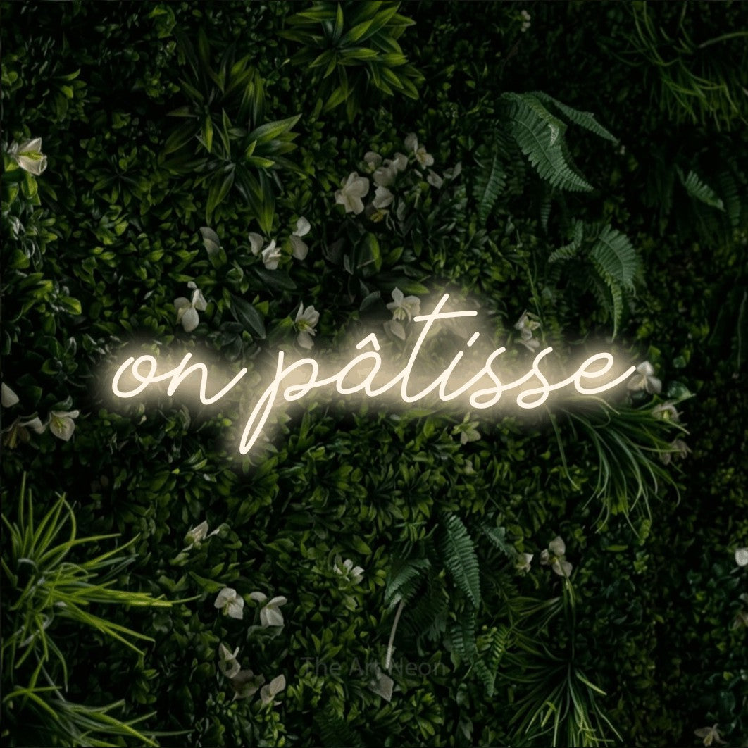 “On pâtisse” LED neon sign in warm white, with cursive lettering, shown against a dense green plant wall.