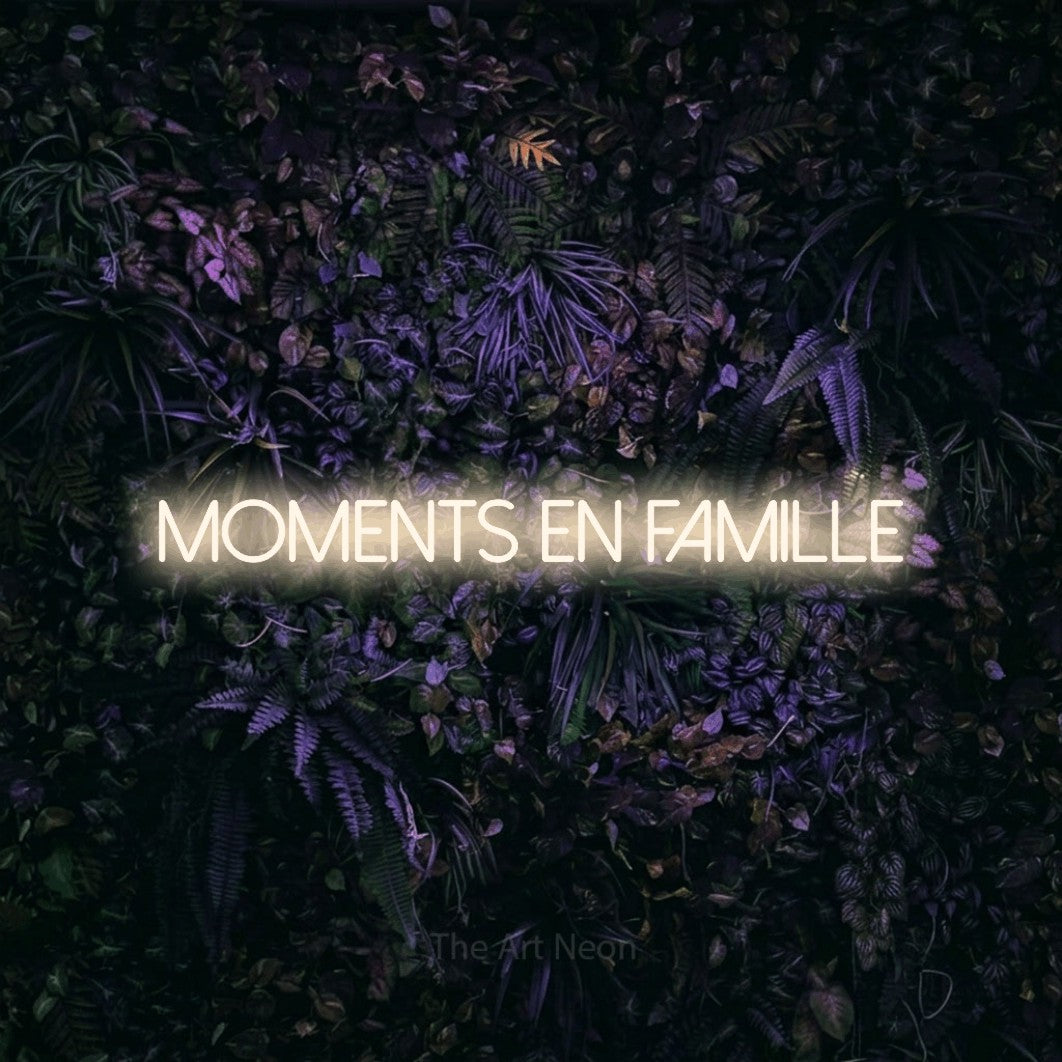 “Moments en famille” LED neon sign in warm white, with thin sans-serif lettering, shown against a dark wall with purple flowers.