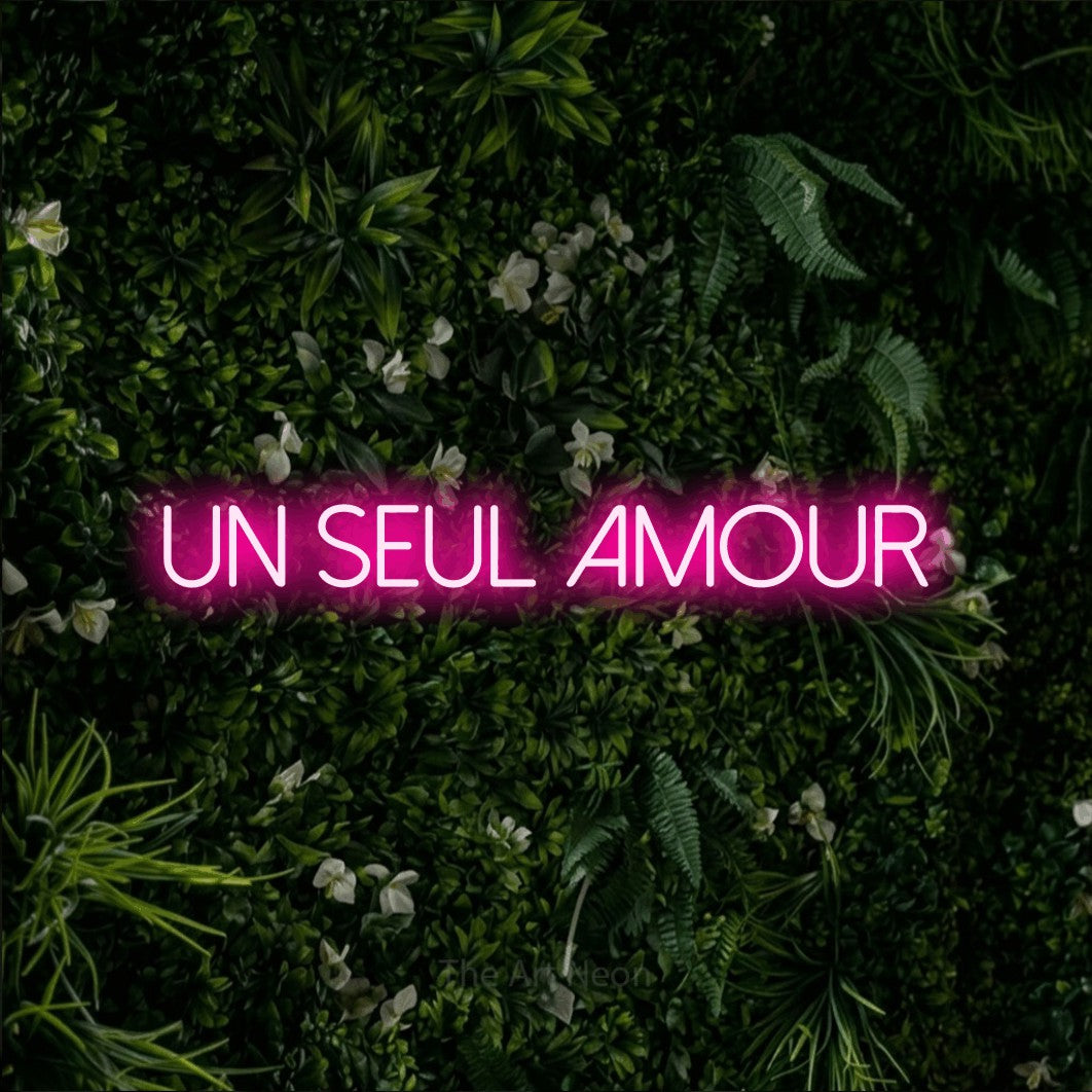 “Un seul amour” LED neon sign in hot pink, with thin sans-serif lettering, shown against a dense green plant wall.