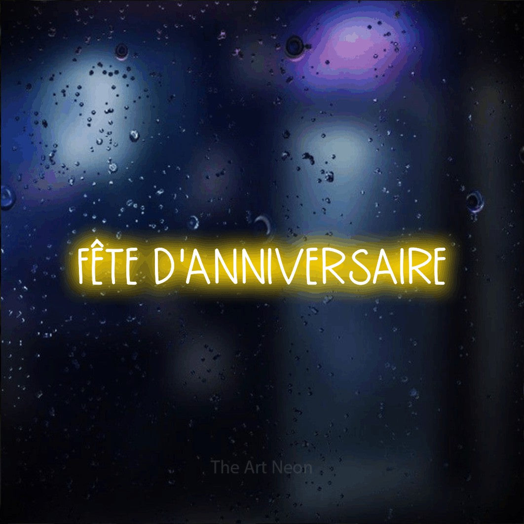 “Fête d'anniversaire” LED neon sign in yellow, with narrow hand-drawn lettering, shown against glass with blue and purple reflections.