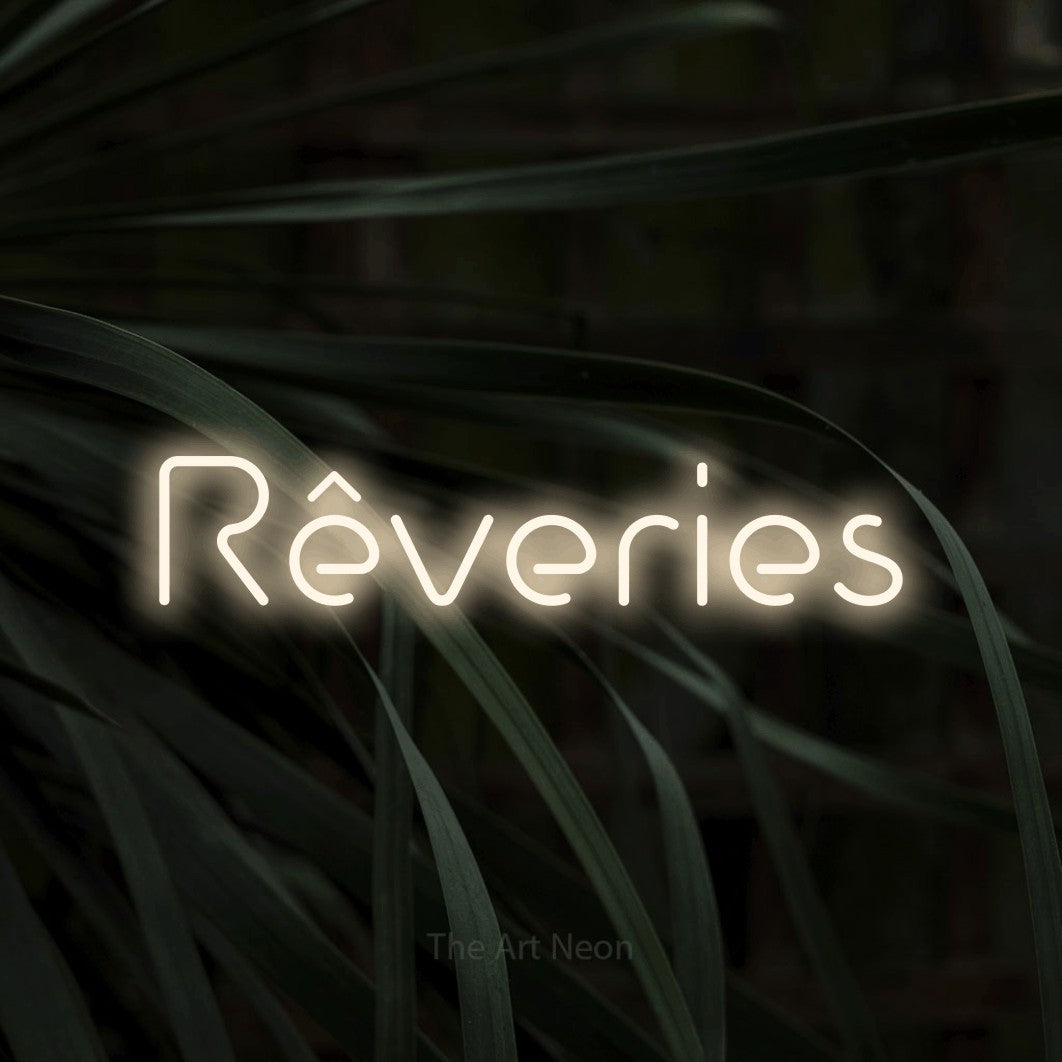 “Rêveries” LED neon sign in warm white, with stylised geometric lettering, shown against a close-up of dark foliage.