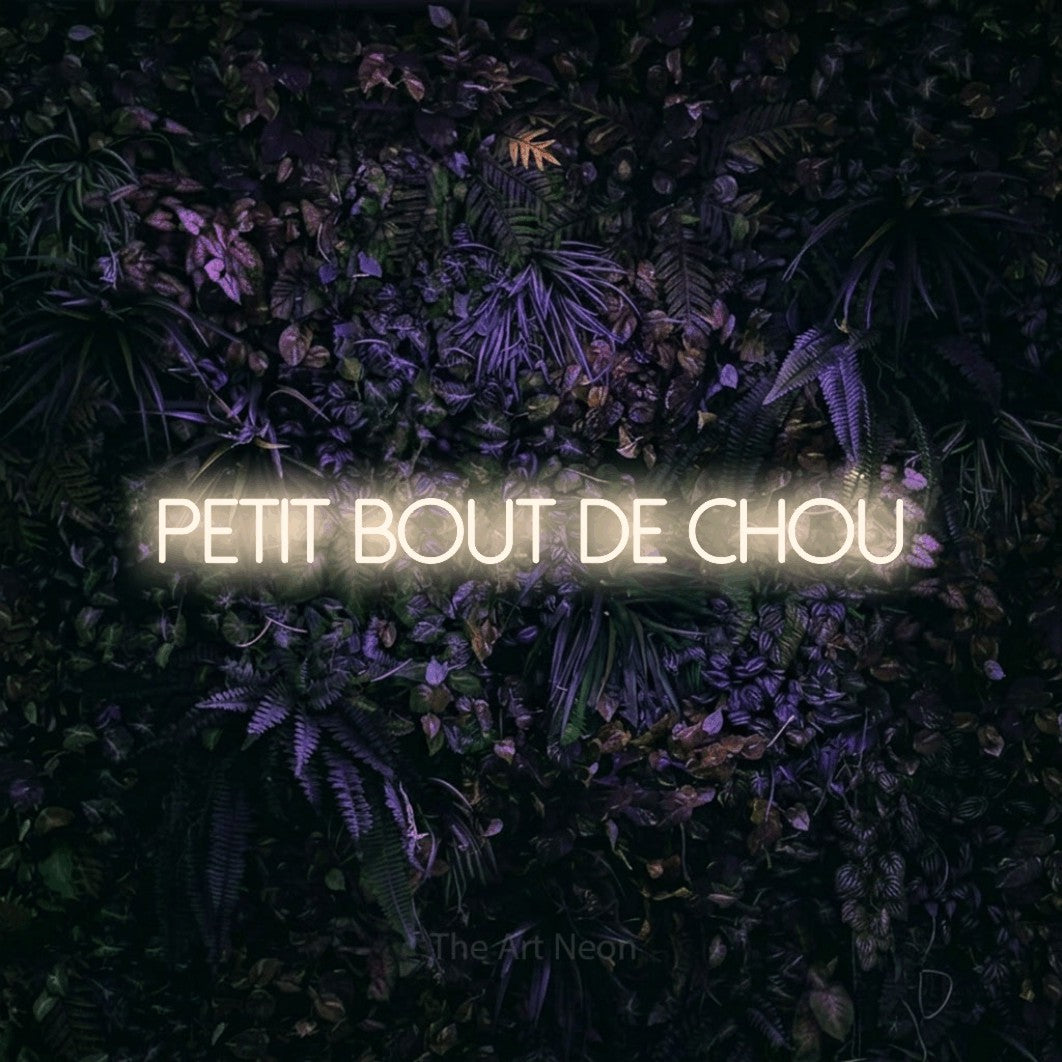 Lettrage en néon LED « Petit bout de chou » en blanc chaud, en lettres fines sans empattement, présenté sur un mur sombre aux fleurs violettes, pour une chambre d’enfant.