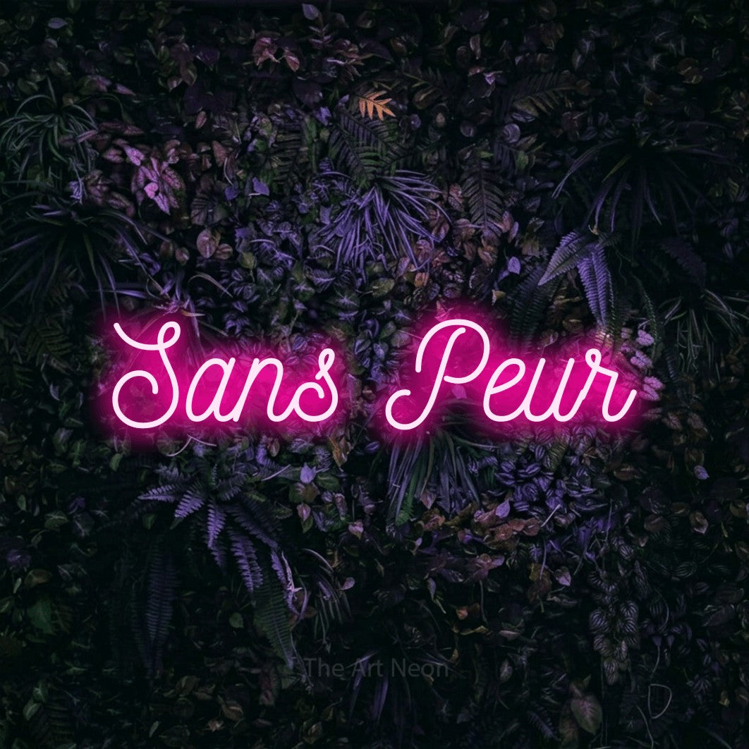“Sans peur” LED neon sign in hot pink, with ornate cursive lettering, shown against a dark wall with purple flowers.