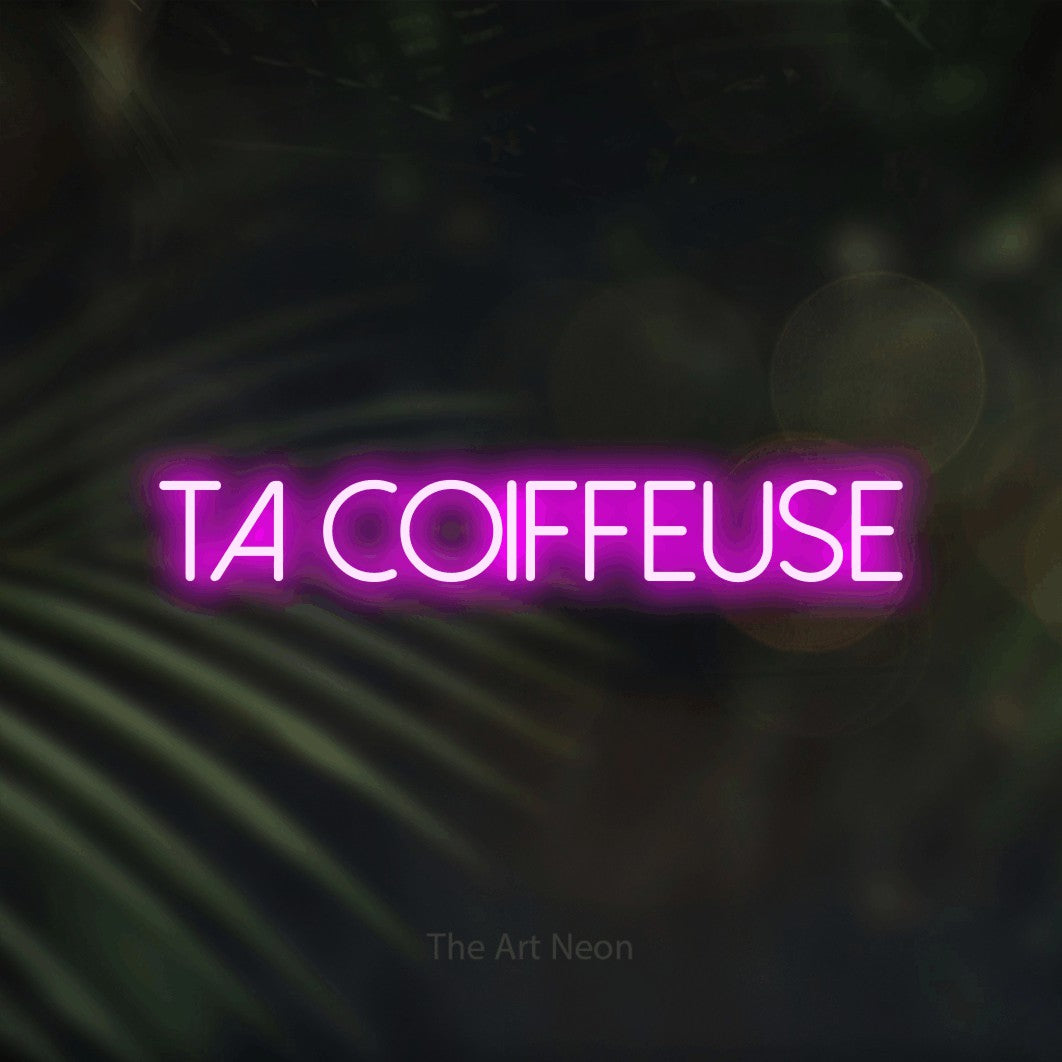 “Ta coiffeuse” LED neon sign in pink, with thin sans-serif lettering, shown against a blurred palm and bokeh-light backdrop.