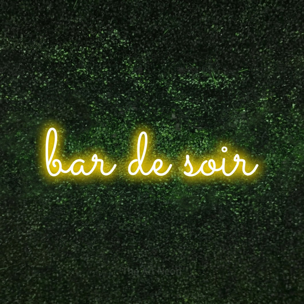 “Bar de soir” LED neon sign in yellow, with looped cursive lettering, shown against a textured green hedge.