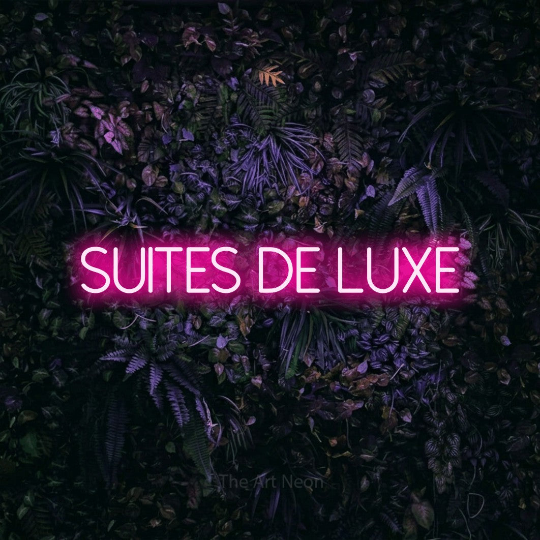“Suites de luxe” LED neon sign in hot pink, with thin sans-serif lettering, shown against a dark wall with purple flowers.