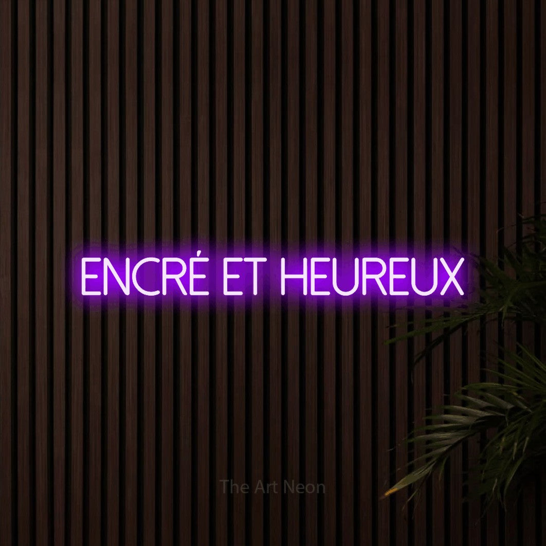 “Encré et heureux” LED neon sign in purple, with thin sans-serif lettering, shown against dark wooden wall slats.