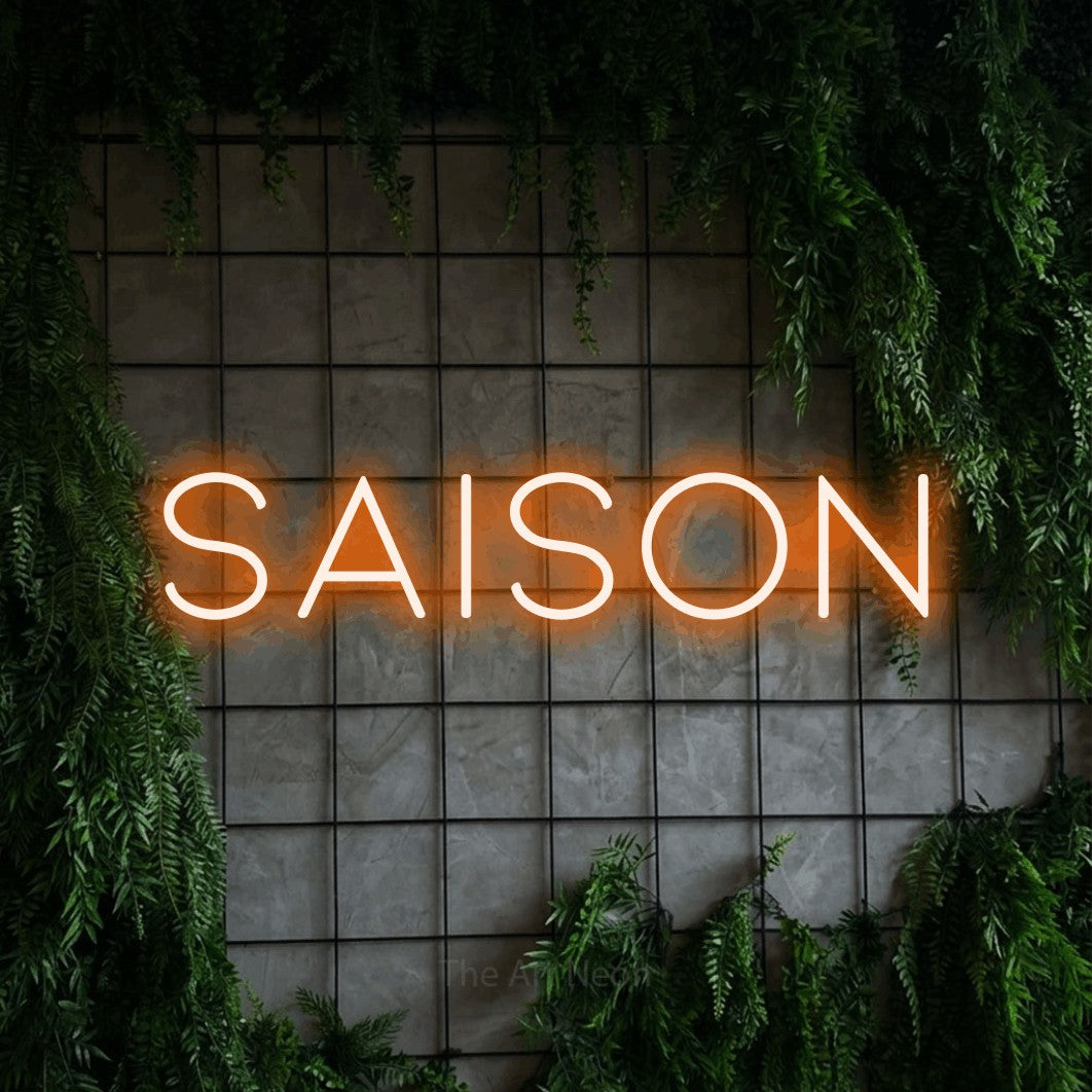 “Saison” LED neon sign in orange, with thin, widely spaced lettering, shown against an ivy-covered concrete grid.