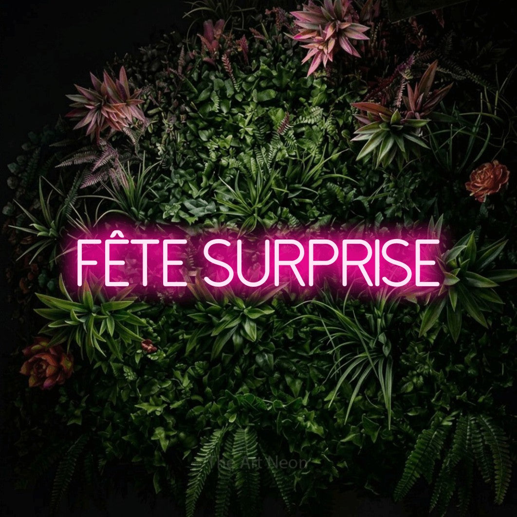 “Fête surprise” LED neon sign in hot pink, with thin sans-serif lettering, shown against a flowering plant wall.