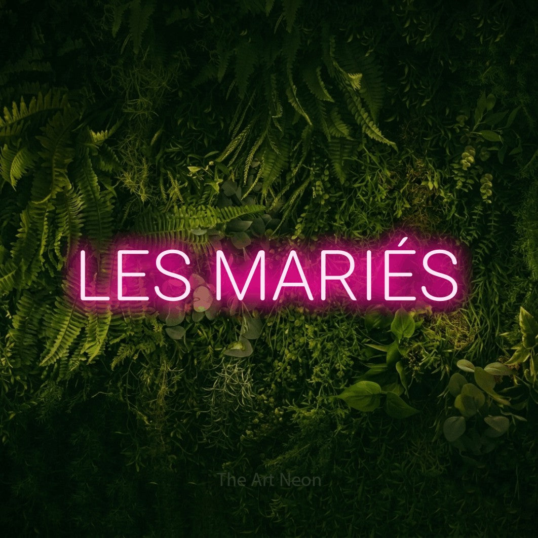 Composition en néon LED « Les mariés » en rose vif, en lettres sans empattement arrondies, mise en valeur sur un fond sombre de mousse et de forêt, pour une réception de mariage.