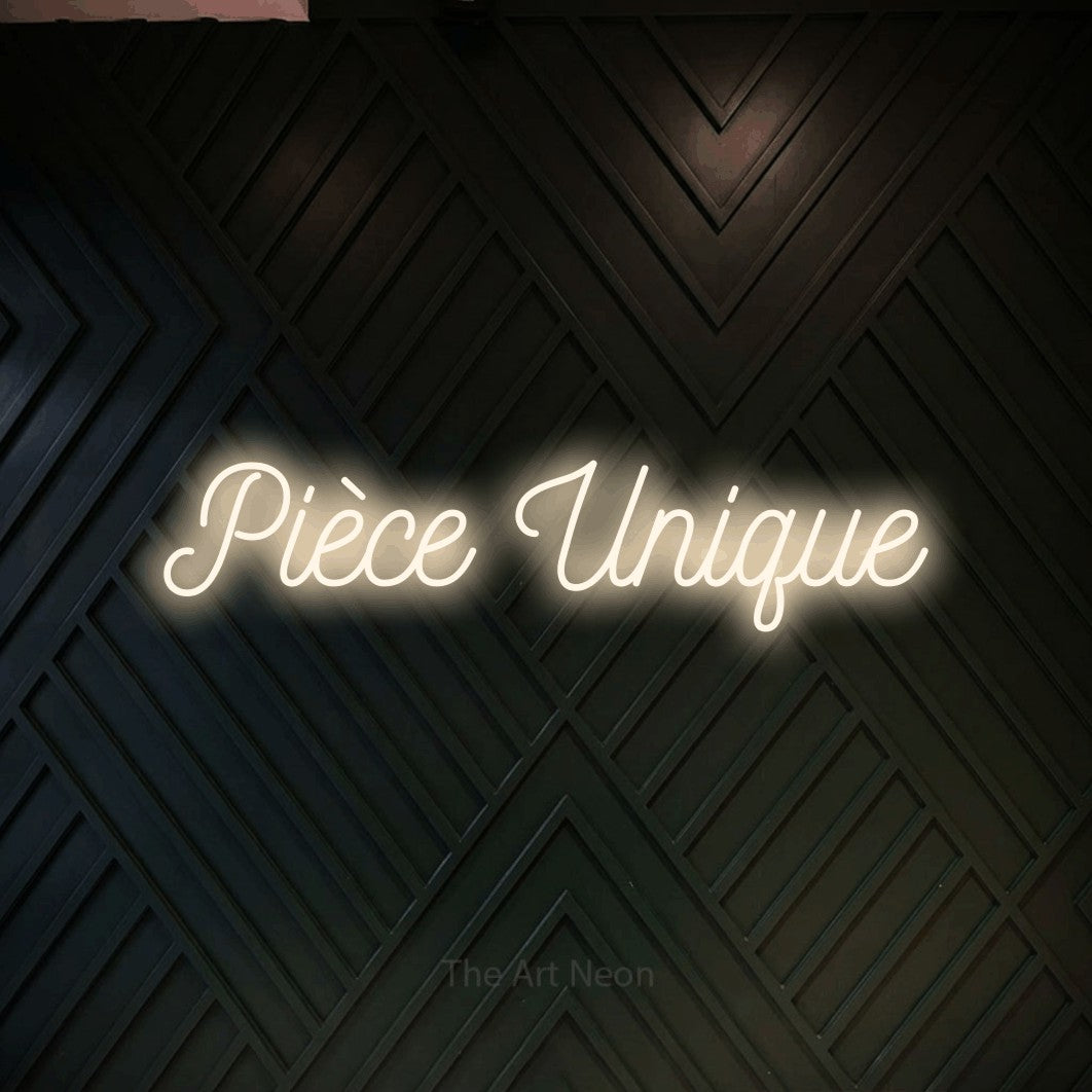“Pièce unique” LED neon sign in warm white, with ornate cursive lettering, shown against a black chevron-patterned wood wall.