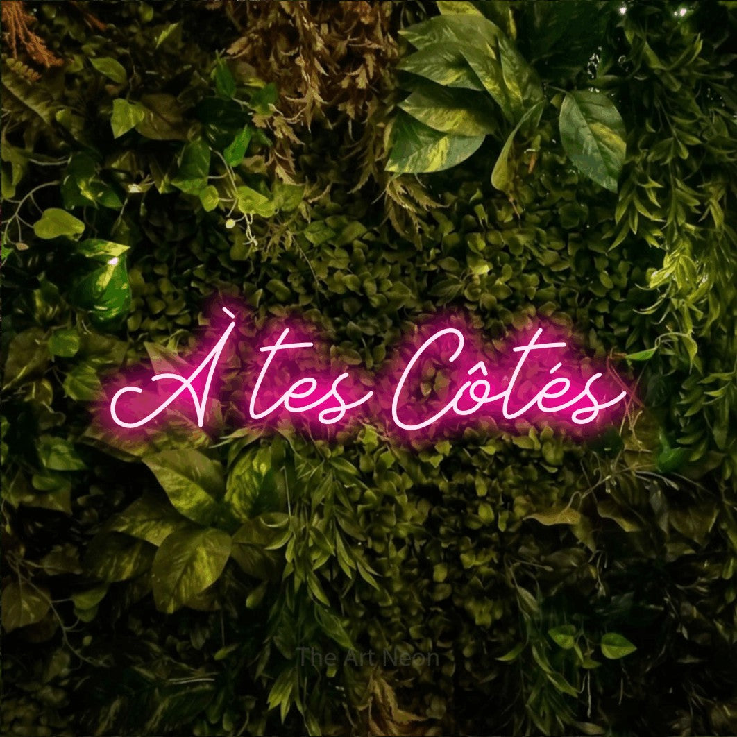 “À tes côtés” LED neon sign in hot pink, with cursive lettering, shown against bright green and yellow foliage.