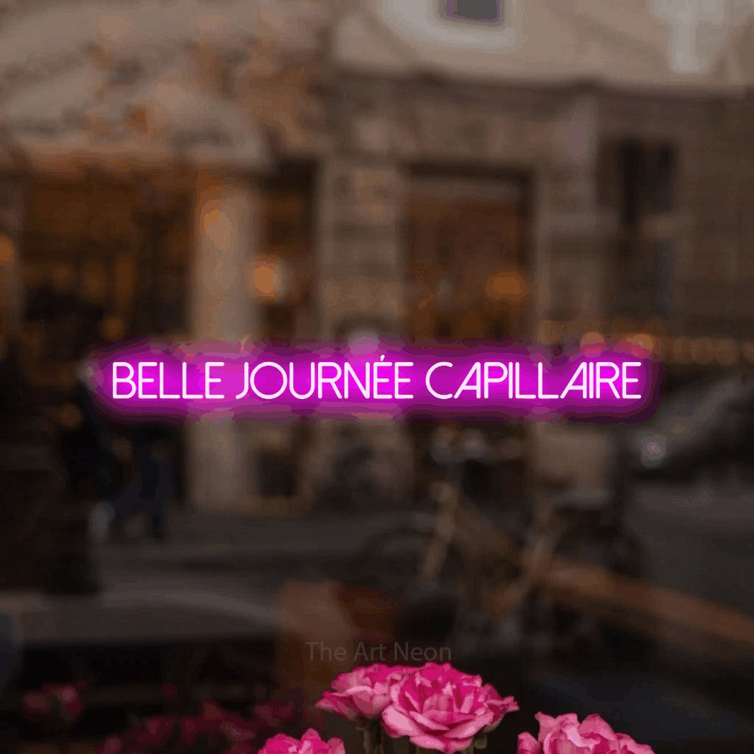 “Belle journée capillaire” LED neon sign in pink, with thin sans-serif lettering, shown against a blurred café-bar interior.