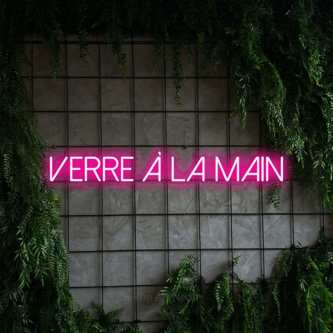 “Verre à la main” LED neon sign in hot pink, with thin sans-serif lettering, shown against an ivy-covered concrete grid.