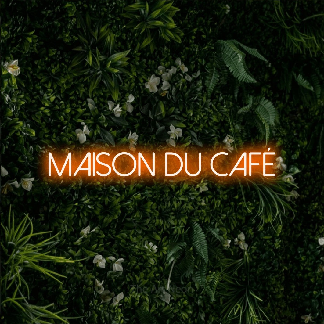 “Maison du café” LED neon sign in orange, with thin sans-serif lettering, shown against a dense green plant wall.