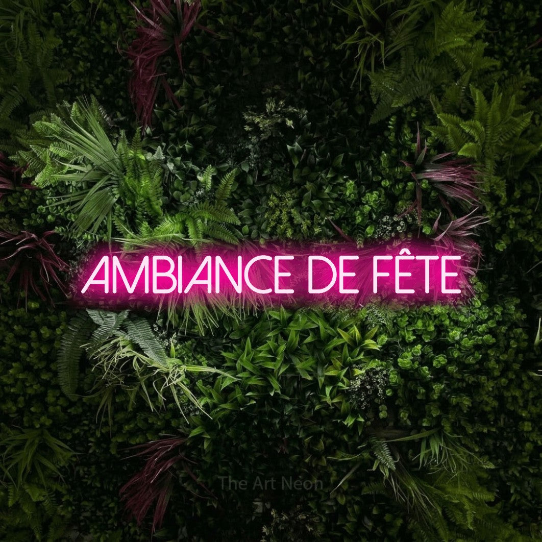 “Ambiance de fête” LED neon sign in hot pink, with thin sans-serif lettering, shown against a plant wall with pink flowers.