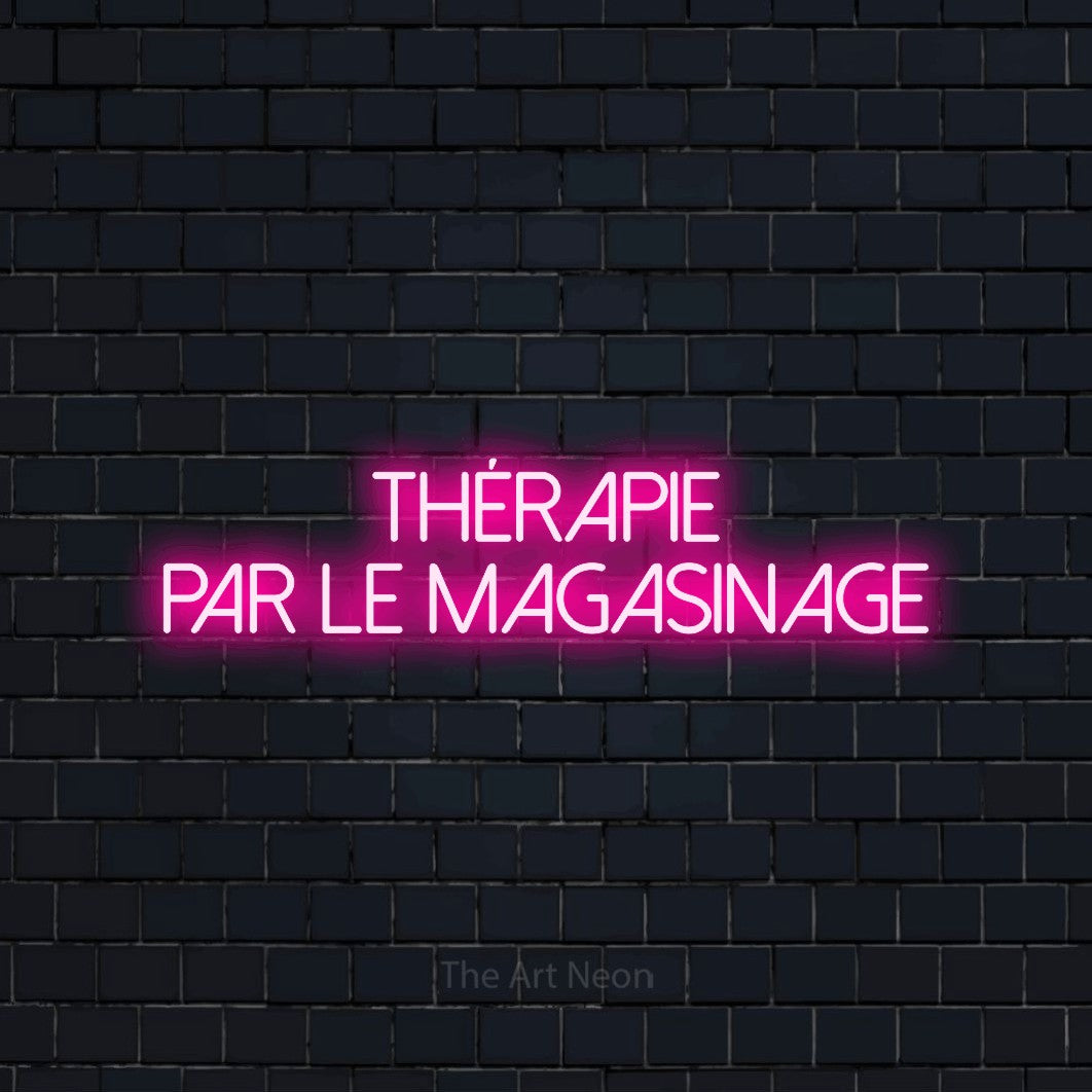 “Thérapie par le magasinage” LED neon sign in hot pink, with thin sans-serif lettering across 2 lines, shown against a dark navy brick wall.