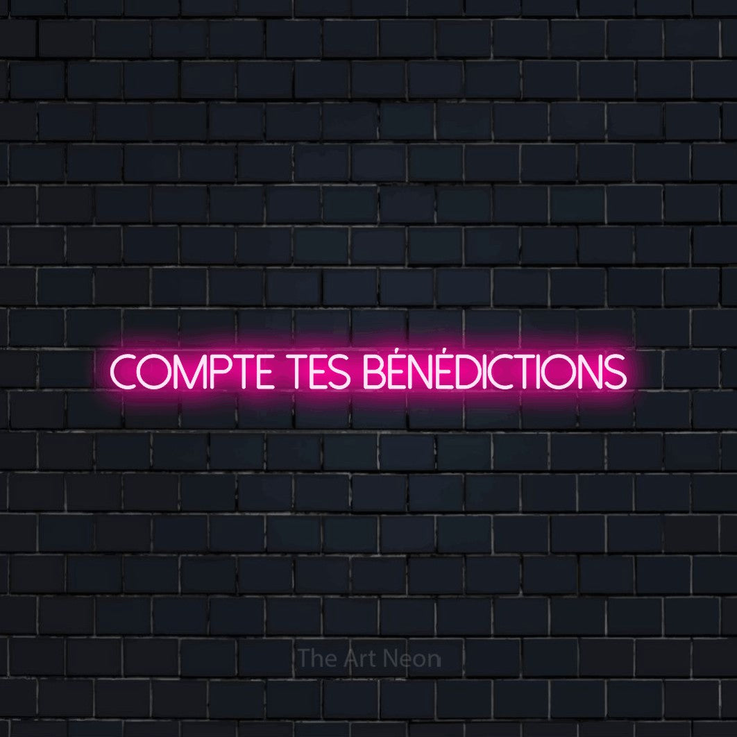 “Compte tes bénédictions” LED neon sign in hot pink, with thin sans-serif lettering, shown against a dark navy brick wall.