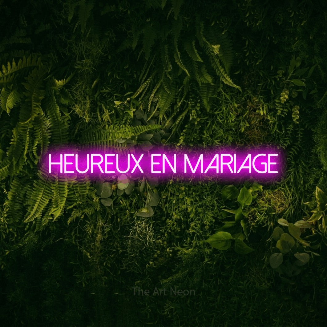 “Heureux en mariage” LED neon sign in pink, with thin sans-serif lettering, shown against a dark moss and forest backdrop.