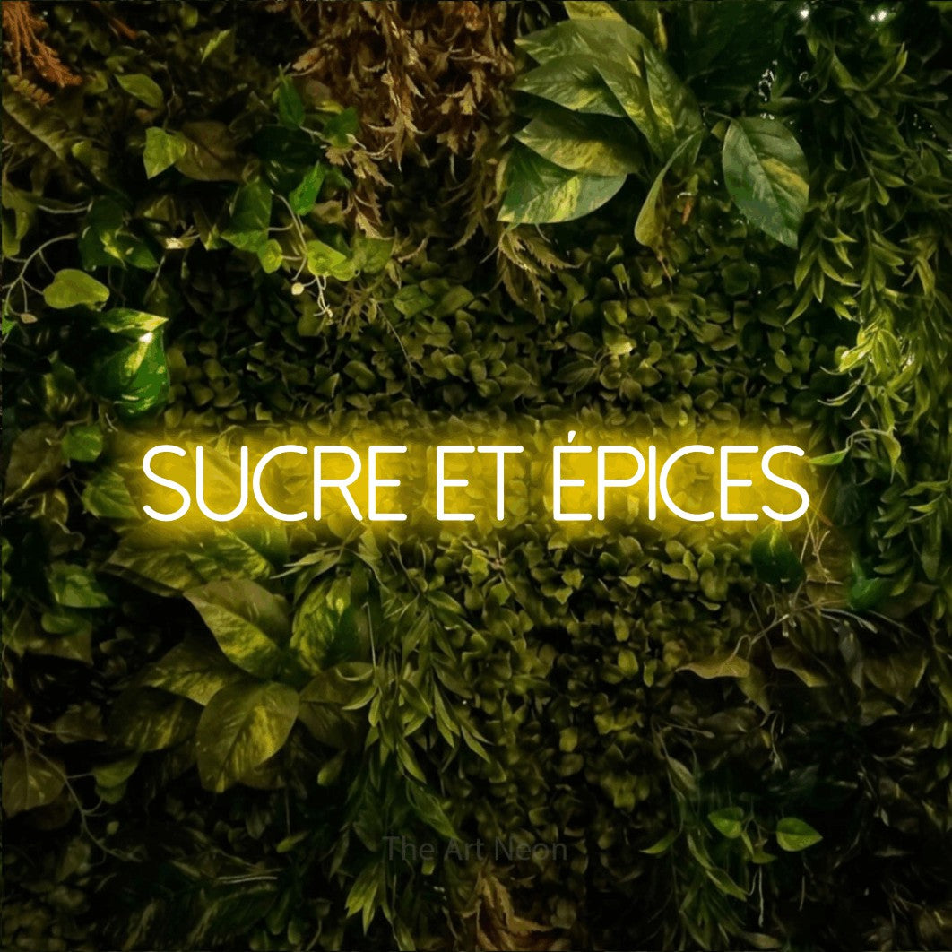 “Sucre et épices” LED neon sign in yellow, with thin sans-serif lettering, shown against bright green and yellow foliage.
