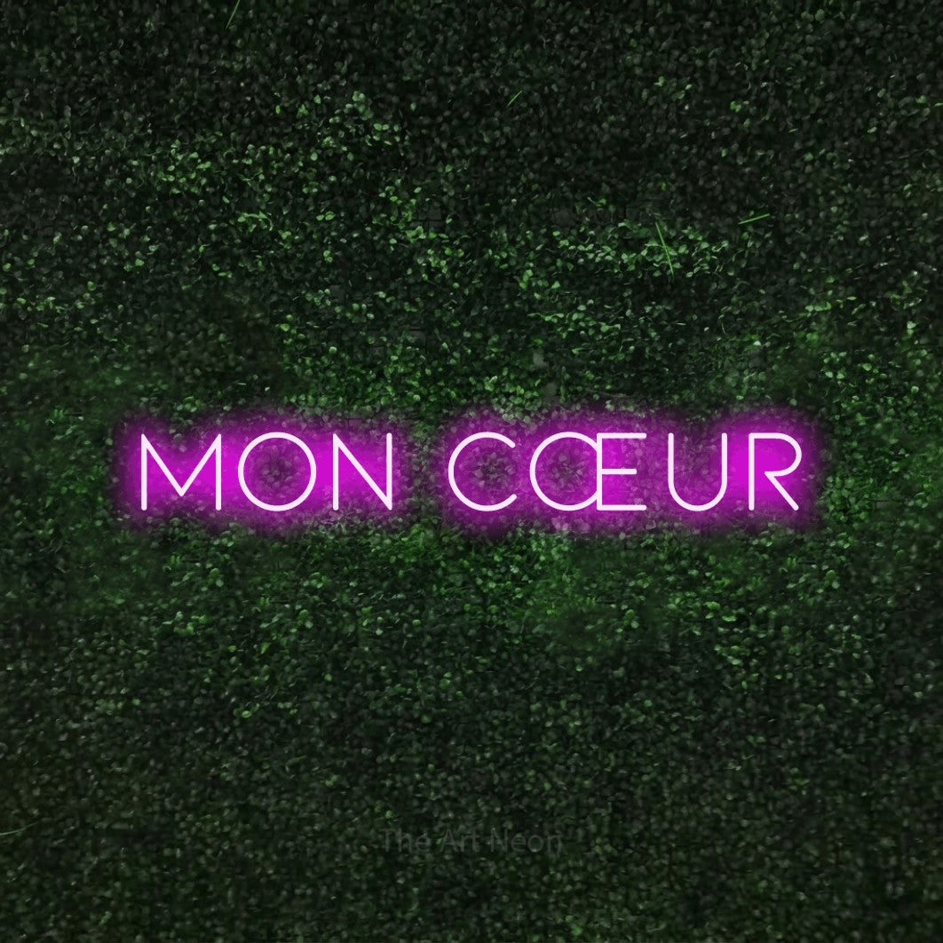 “Mon cœur” LED neon sign in pink, with thin, widely spaced lettering, shown against a textured green hedge.