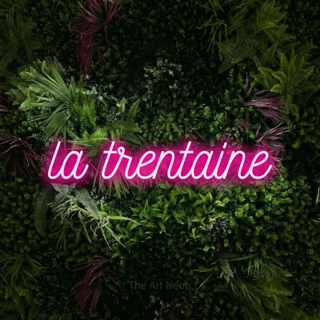 “La trentaine” LED neon sign in hot pink, with ornate cursive lettering, shown against a plant wall with pink flowers.