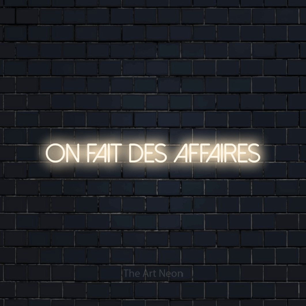 “On fait des affaires” LED neon sign in warm white, with thin sans-serif lettering, shown against a dark navy brick wall.