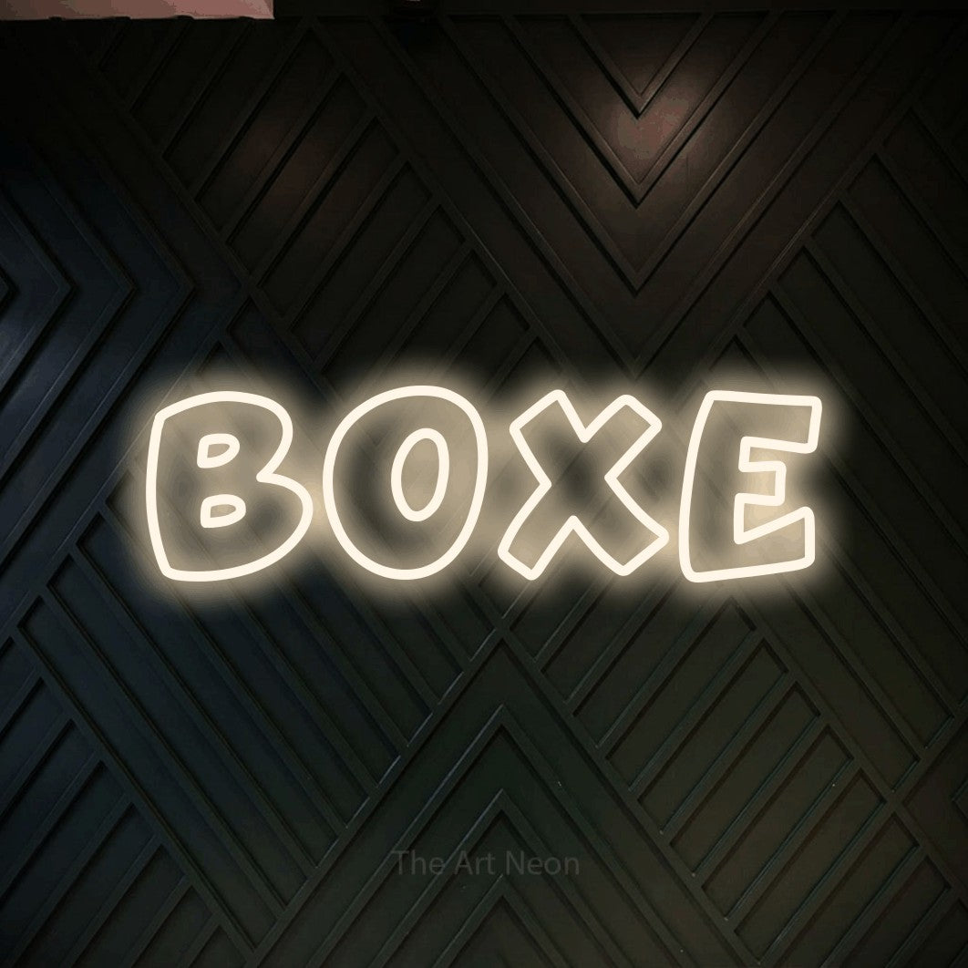 Néon LED « Boxe » en blanc chaud, en lettres arrondies dessinées en contour, mis en scène sur un mur en bois noir à motif chevron, pour un espace d’entraînement.