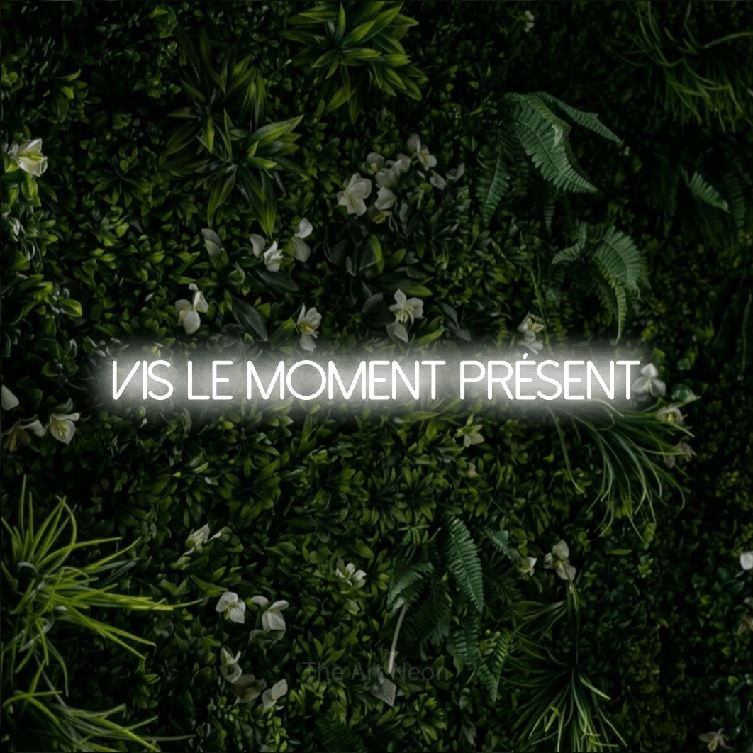 “Vis le moment présent” LED neon sign in cool white, with thin sans-serif lettering, shown against a dense green plant wall.