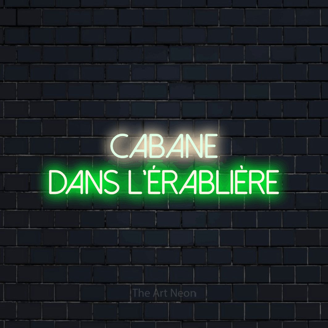 “Cabane dans l'érablière” LED neon sign in warm white on the upper line and green on the lower line, with thin sans-serif lettering across 2 lines, shown against a dark navy brick wall.
