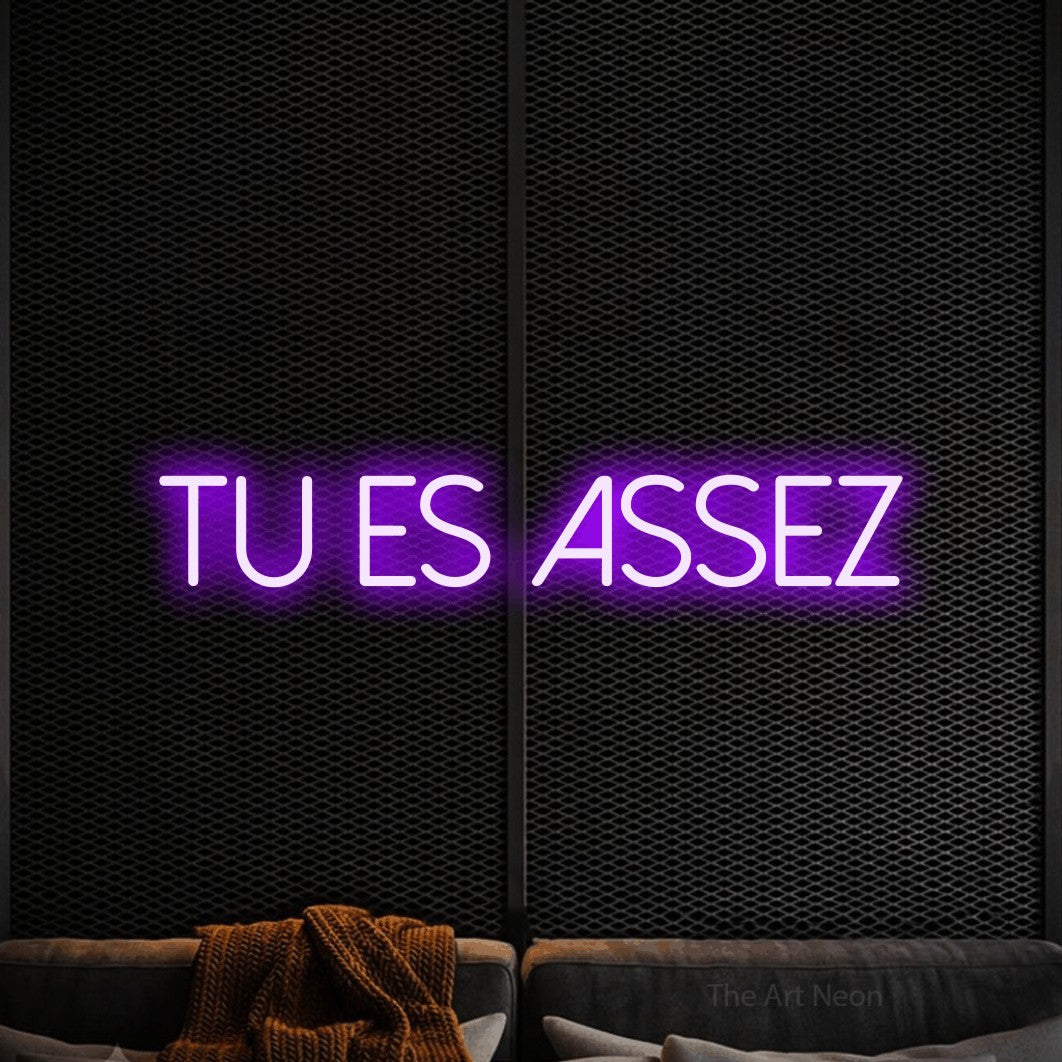 “Tu es assez” LED neon sign in purple, with thin sans-serif lettering, shown against a modern dark-toned seating area.