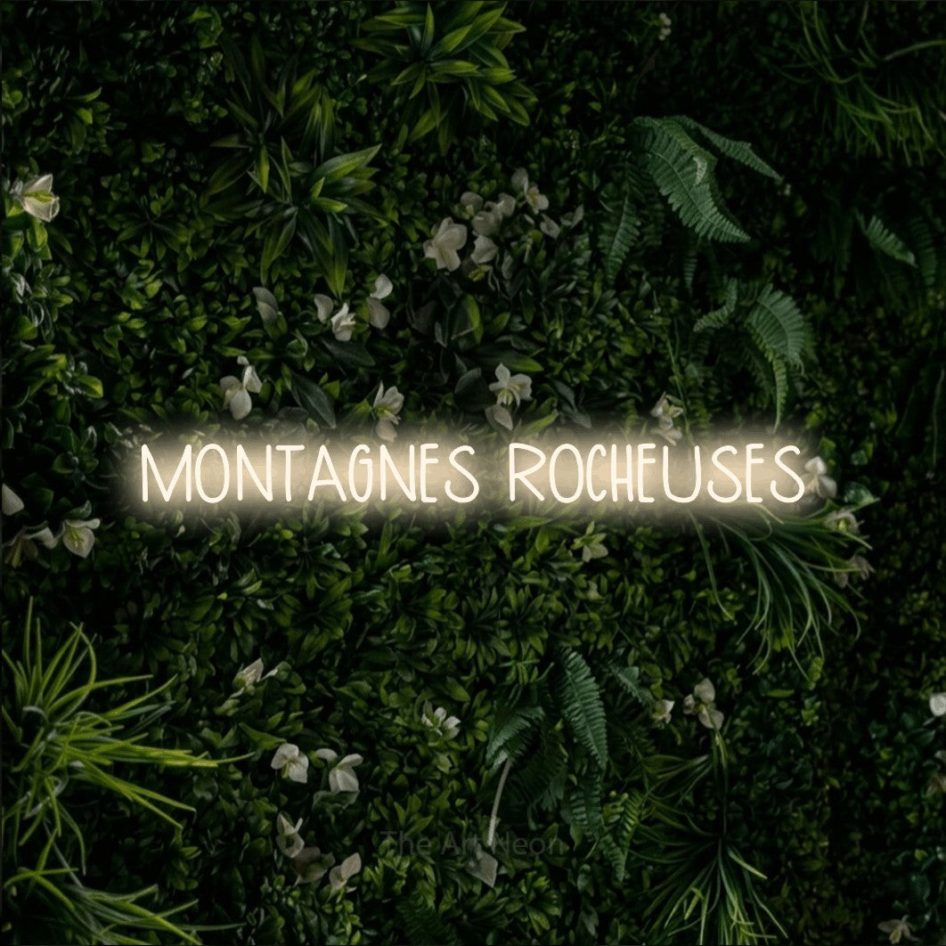 “Montagnes rocheuses” LED neon sign in warm white, with narrow hand-drawn lettering, shown against a dense green plant wall.