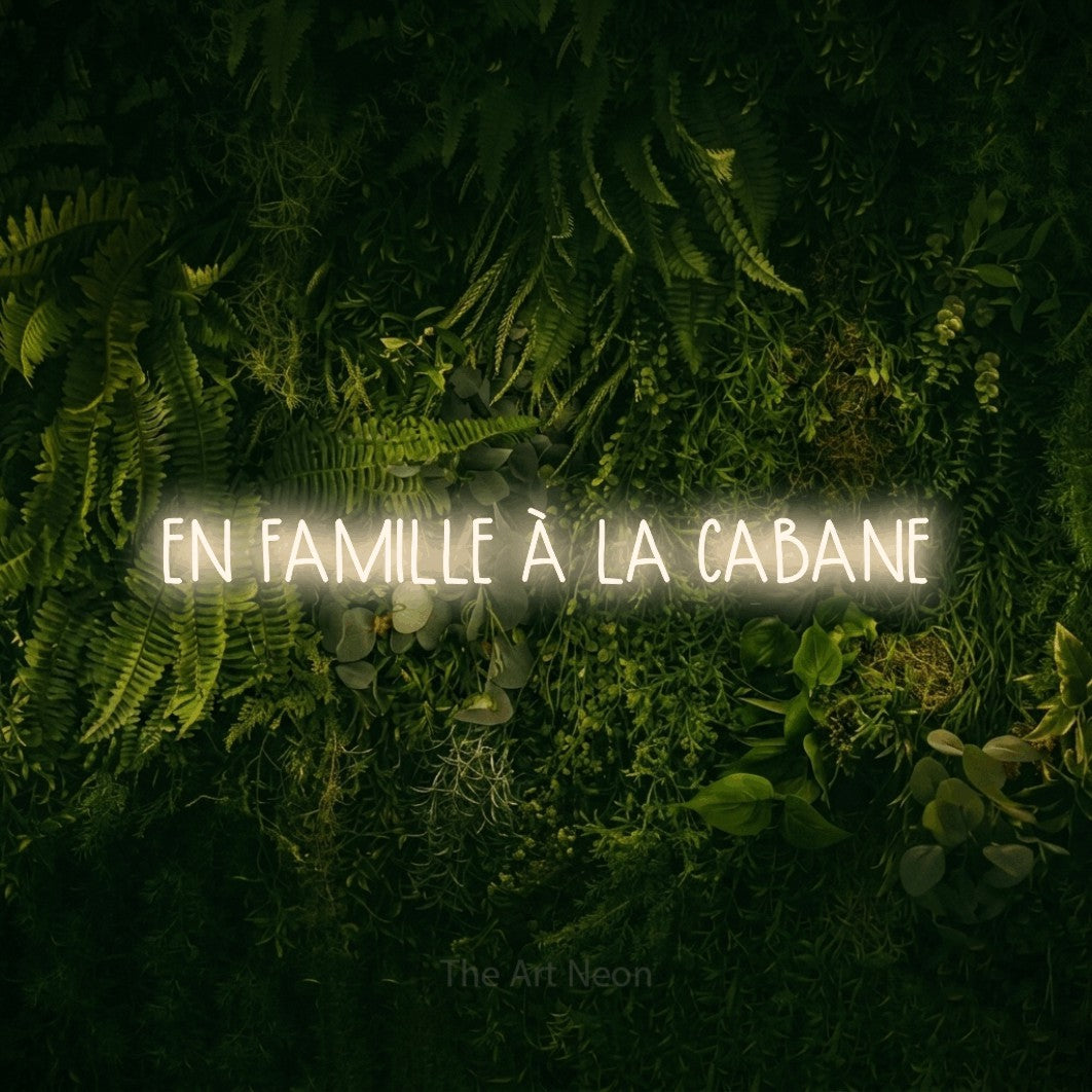 “En famille à la cabane” LED neon sign in warm white, with narrow hand-drawn lettering, shown against a dark moss and forest backdrop.