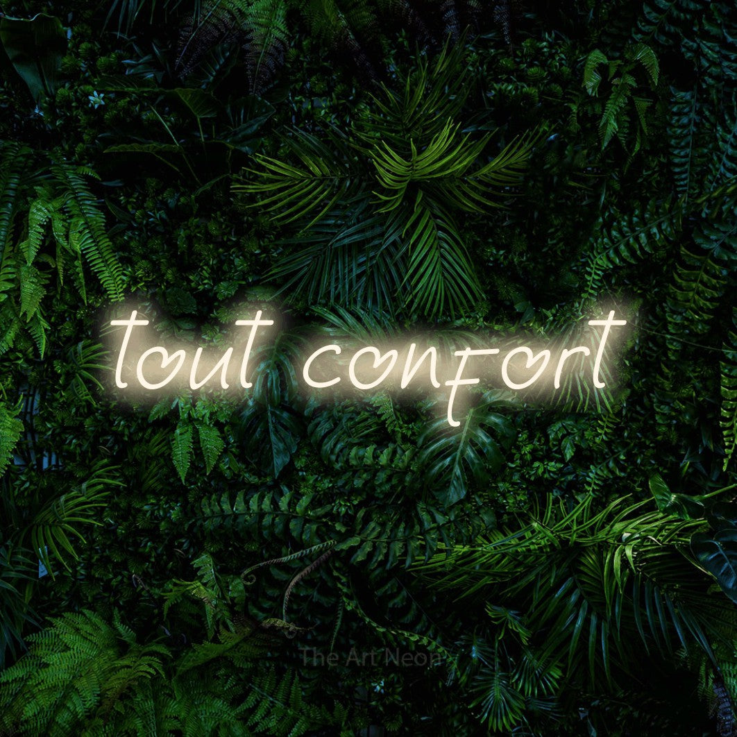 “Tout confort” LED neon sign in warm white, with slanted handwritten lettering, shown against dense tropical foliage.