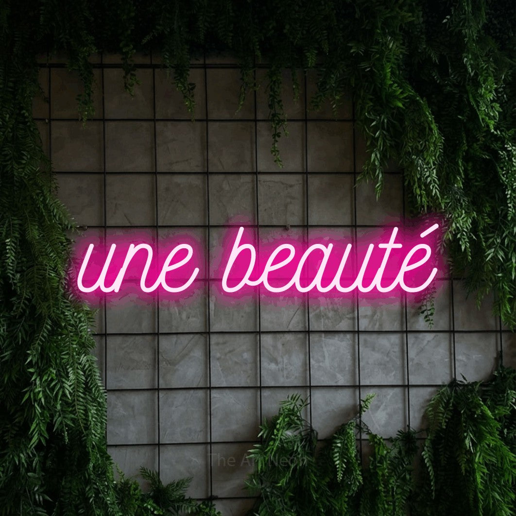 “Une beauté” LED neon sign in hot pink, with ornate cursive lettering, shown against an ivy-covered concrete grid.