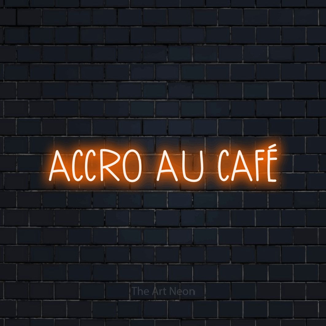 “Accro au café” LED neon sign in orange, with narrow hand-drawn lettering, shown against a dark navy brick wall.