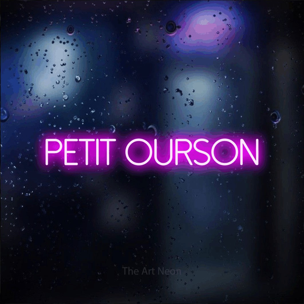 “Petit ourson” LED neon sign in pink, with thin sans-serif lettering, shown against glass with blue and purple reflections.