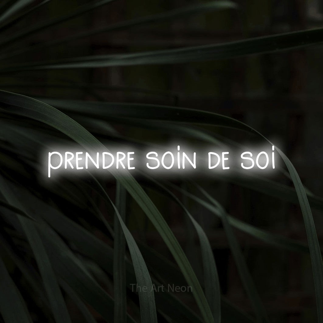 “Prendre soin de soi” LED neon sign in cool white, with narrow hand-drawn lettering, shown against a close-up of dark foliage.