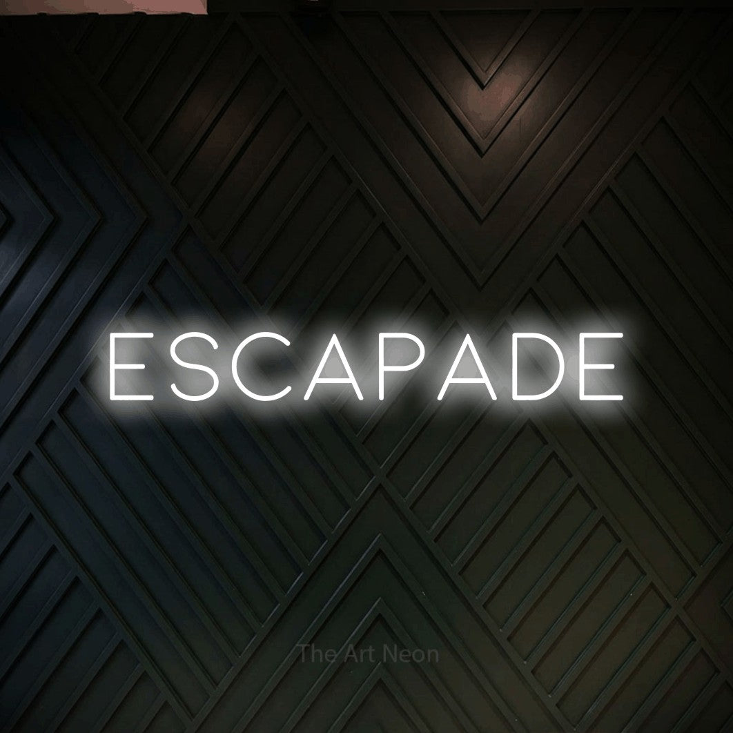 “Escapade” LED neon sign in cool white, with thin, widely spaced lettering, shown against a black chevron-patterned wood wall.