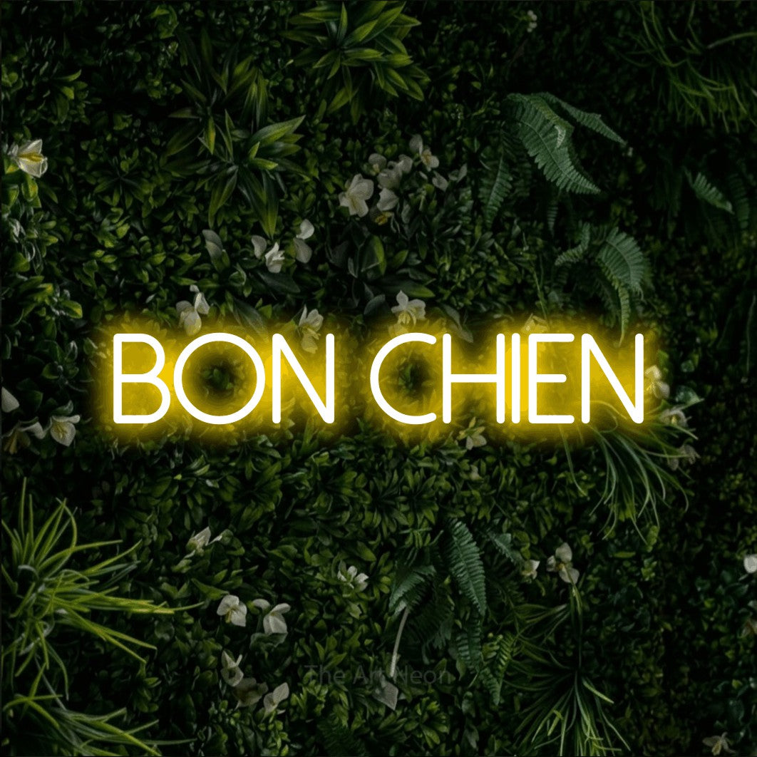 “Bon chien” LED neon sign in yellow, with thin sans-serif lettering, shown against a dense green plant wall.