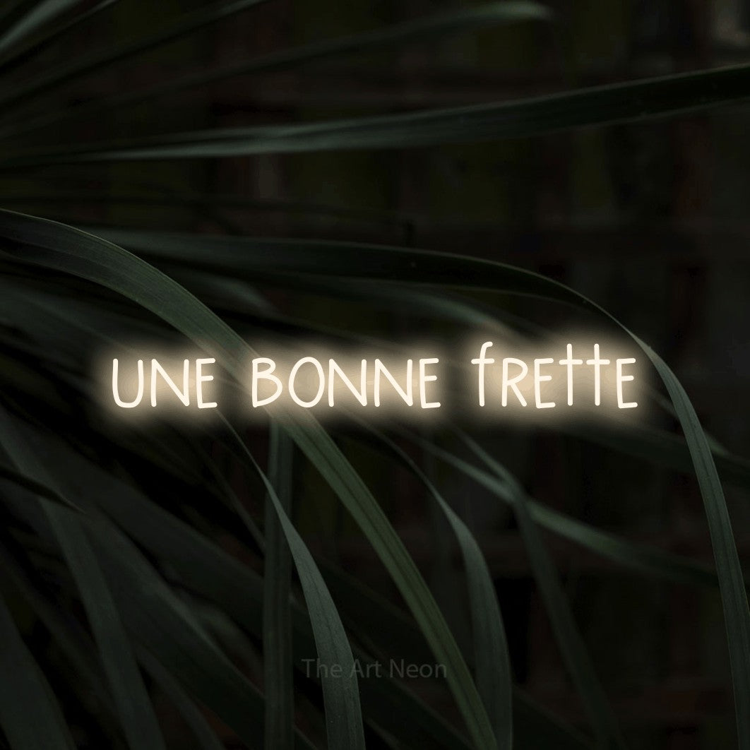 “Une bonne frette” LED neon sign in warm white, with narrow hand-drawn lettering, shown against a close-up of dark foliage.
