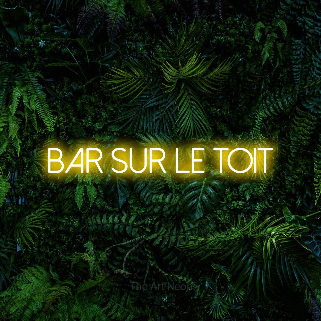 “Bar sur le toit” LED neon sign in yellow, with thin sans-serif lettering, shown against dense tropical foliage.
