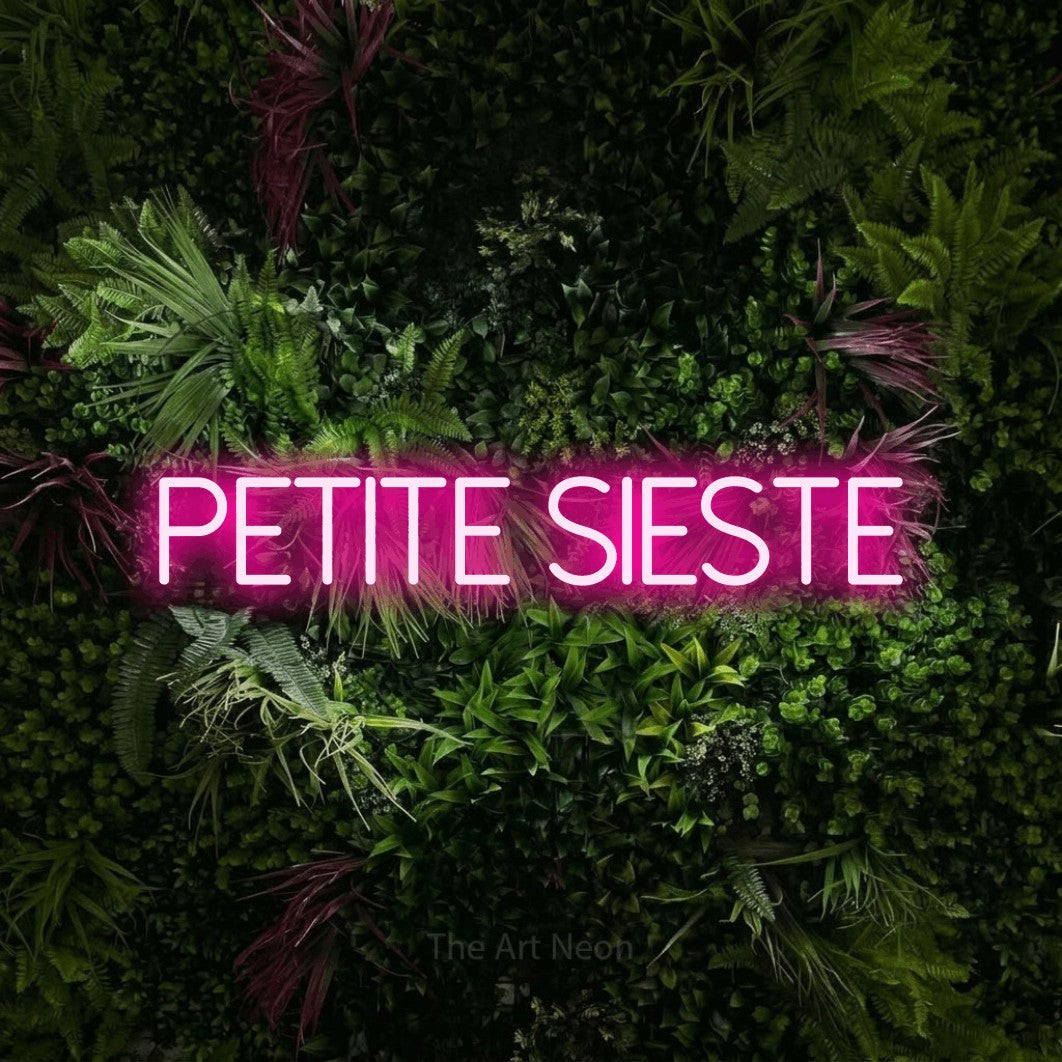 “Petite sieste” LED neon sign in hot pink, with thin sans-serif lettering, shown against a plant wall with pink flowers.
