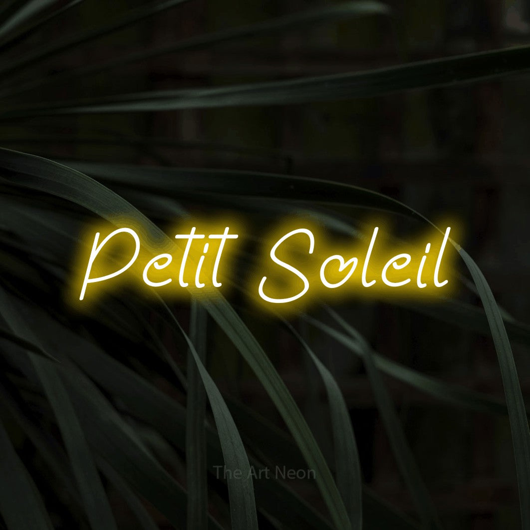 “Petit soleil” LED neon sign in yellow, with slanted handwritten lettering, shown against a close-up of dark foliage.