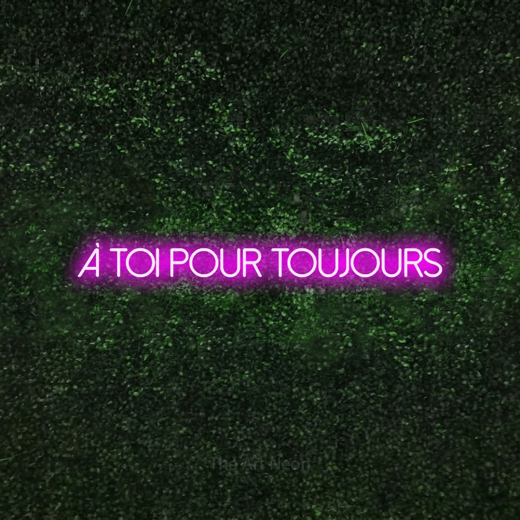 “À toi pour toujours” LED neon sign in pink, with thin sans-serif lettering, shown against a textured green hedge.