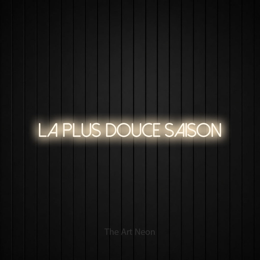 “La plus douce saison” LED neon sign in warm white, with thin sans-serif lettering, shown against black vertical wall slats.