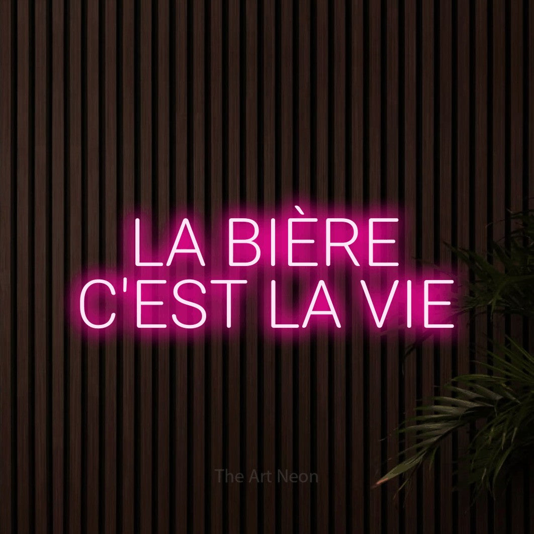 Lettrage en néon LED « La bière c'est la vie » sur deux lignes en rose vif, en lettres sans empattement arrondies, présenté sur un mur de lattes en bois foncé, pour une microbrasserie.