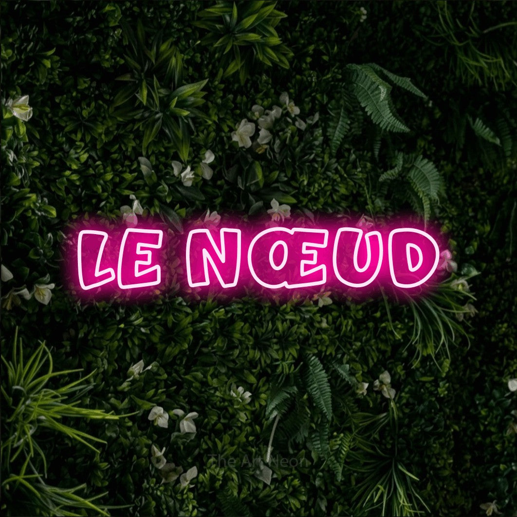 “Le nœud” LED neon sign in hot pink, with rounded outline lettering, shown against a dense green plant wall.