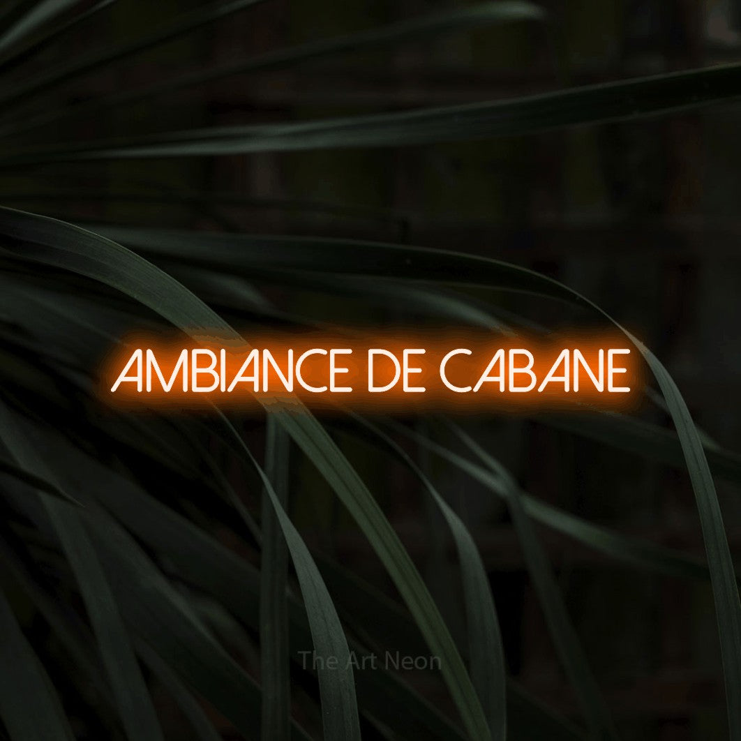 “Ambiance de cabane” LED neon sign in orange, with thin sans-serif lettering, shown against a close-up of dark foliage.